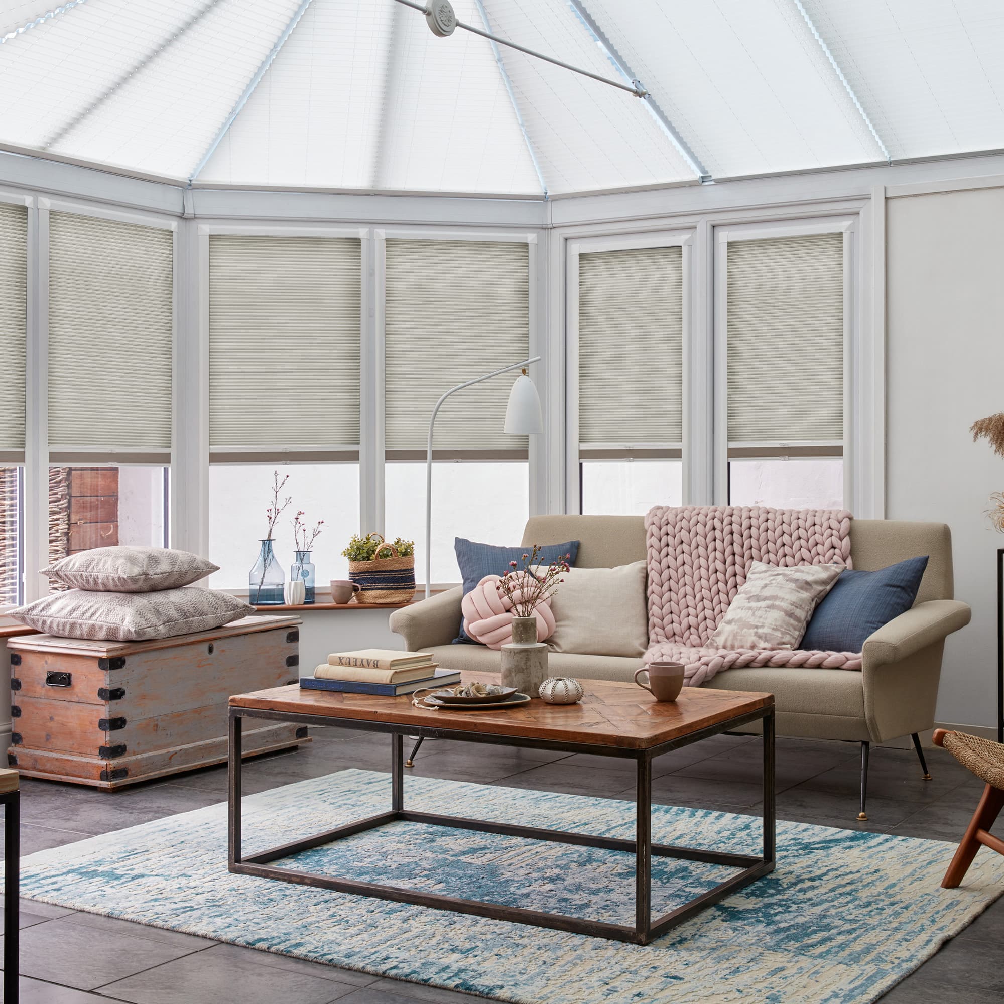 Cellular pleated blinds drawn down over tall conservatory windows, lightly diffusing daylight; beige blinds in a bright sunroom living area with a sofa, coffee table, knit throw, and blue rug.