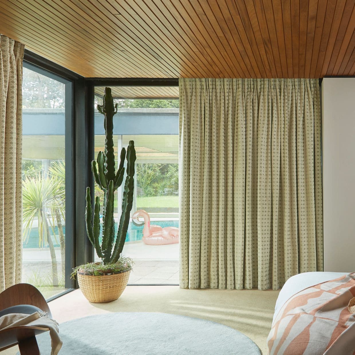 Pleated patterned floor-to-ceiling drapes, drawn closed over the right sliding glass door; filtering daylight. Mid-century bedroom with wood-paneled ceiling, potted cactus by the glass and pool with pink flamingo outside.