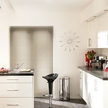 Beige horizontal blinds covering the window, closed and filtering soft daylight; modern white kitchen with black barstool, stainless bin, wall clock, coffee maker and vase of red tulips.