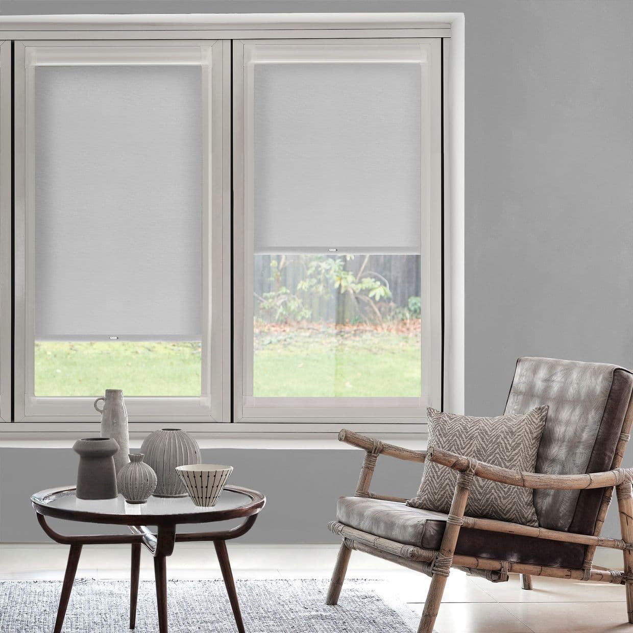 Roller blinds covering two tall windows, partially lowered; filtering soft daylight into a neutral-toned living room with a wooden armchair, patterned cushion, low coffee table and garden view.