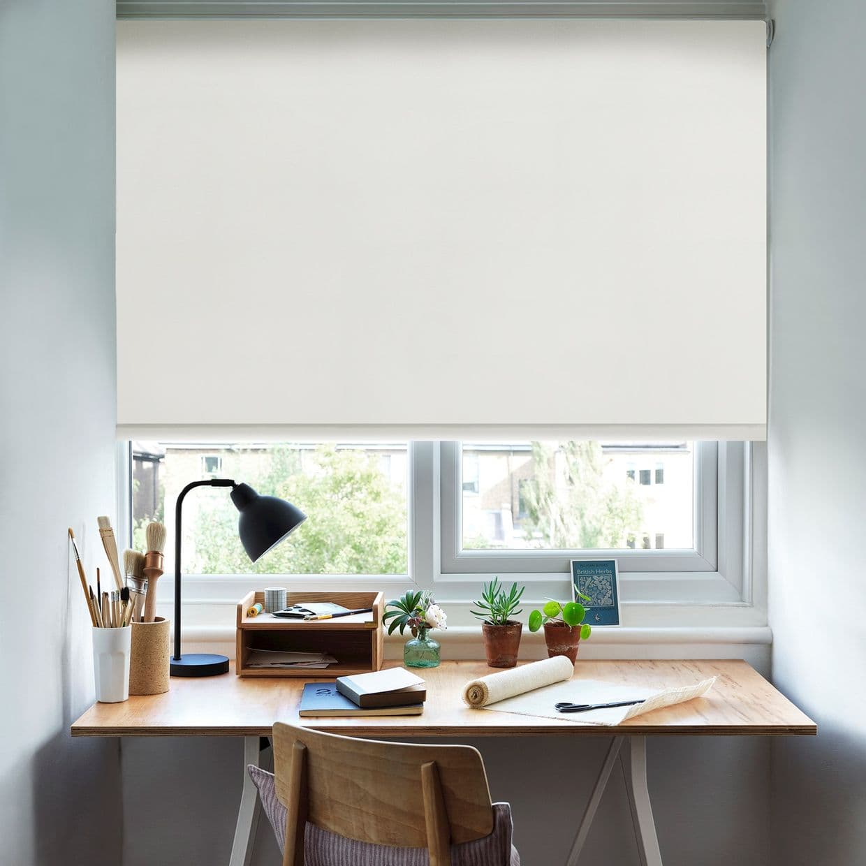 White roller blind lowered near the sill, opaque fabric blocking daylight; wooden desk beneath with lamp, brushes, notebooks and small potted succulents in a compact home study.