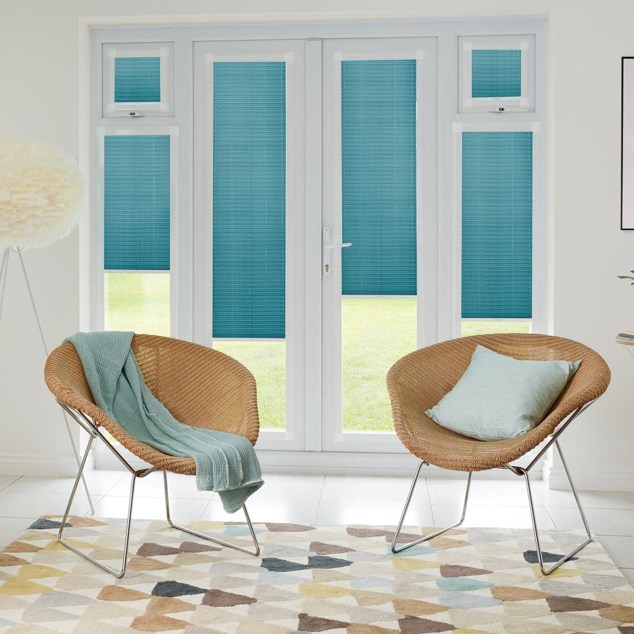 Teal pleated blinds on white French doors and sidelights, lowered and filtering light; modern sitting room with two rattan chairs, pale throw and cushion, geometric rug, view of green lawn.