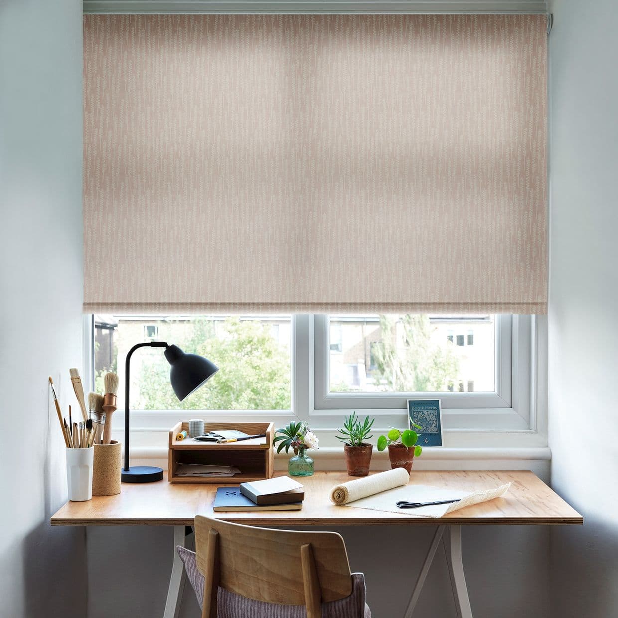 Pale patterned roller blind lowered, gently filtering daylight and covering the window. A wooden desk beneath holds a black lamp, art brushes, small potted plants and notebooks in a bright home office.