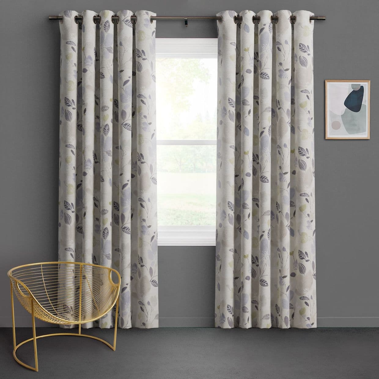 Grommet-top, leaf-patterned curtains hung on a metal rod, parted at the center and filtering soft daylight; framed by grey walls, a gold wire chair and abstract wall art.