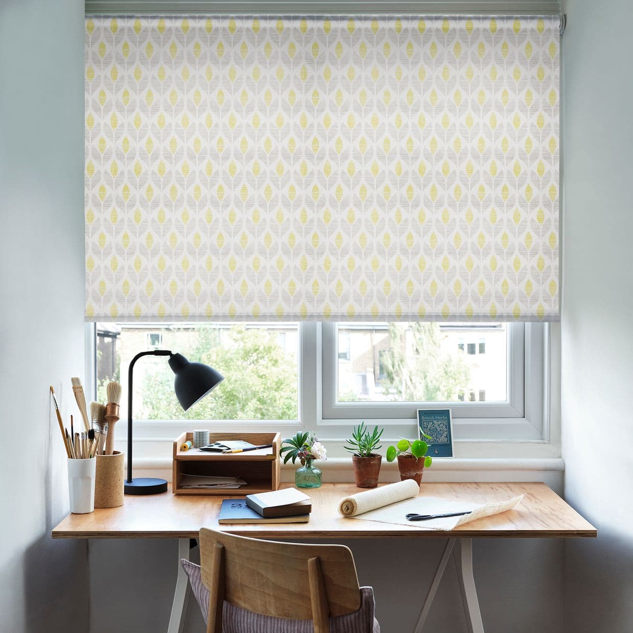 Patterned roller blind lowered over the windowâ€”pale grey fabric with yellow leaf motifâ€”filtering soft daylight; tidy wooden desk beneath holds a lamp, art brushes, notebooks and small potted plants.