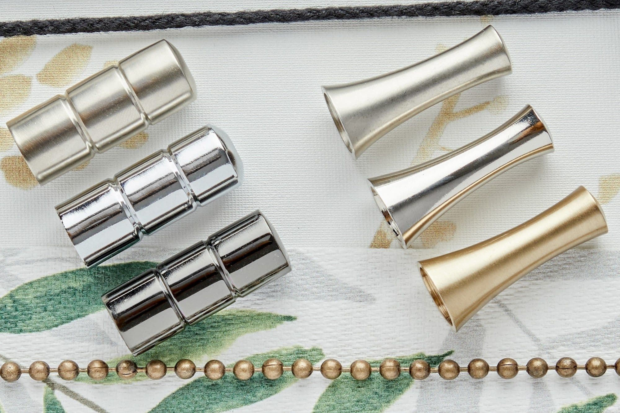 Metal curtain-rod components â€” three segmented chrome connectors (left) and three flared cone finials in silver and gold (right) â€” lie arranged on white leaf-print fabric under soft overhead light.
