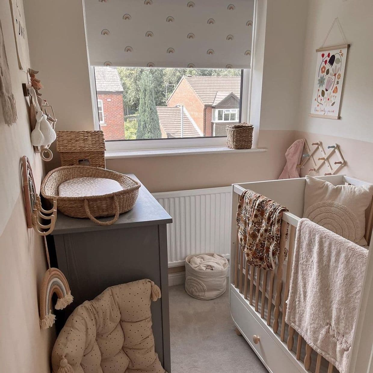 Roller blind with small rainbow print, lowered over window, softly filtering daylight; nursery with crib, changing basket on dresser, neutral decor and outside view of brick houses and trees.