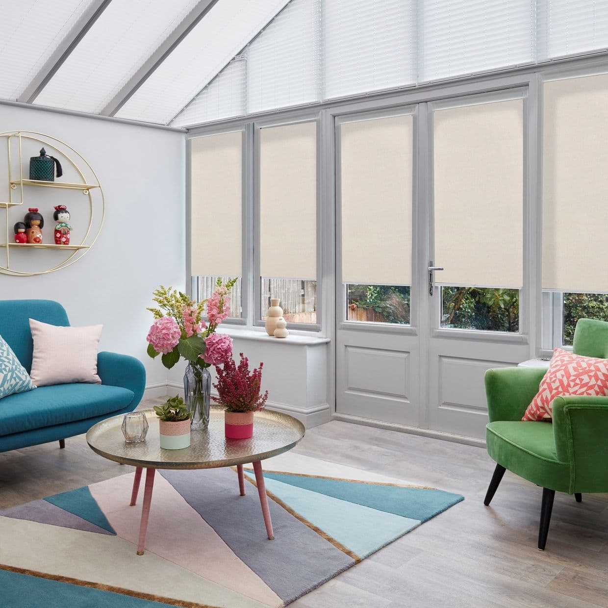Beige roller blinds lowered mid-height across tall glazed doors and sloped roof windows, partially closed and filtering daylight; conservatory-style living room with teal sofa, green armchair, round coffee table, rug.