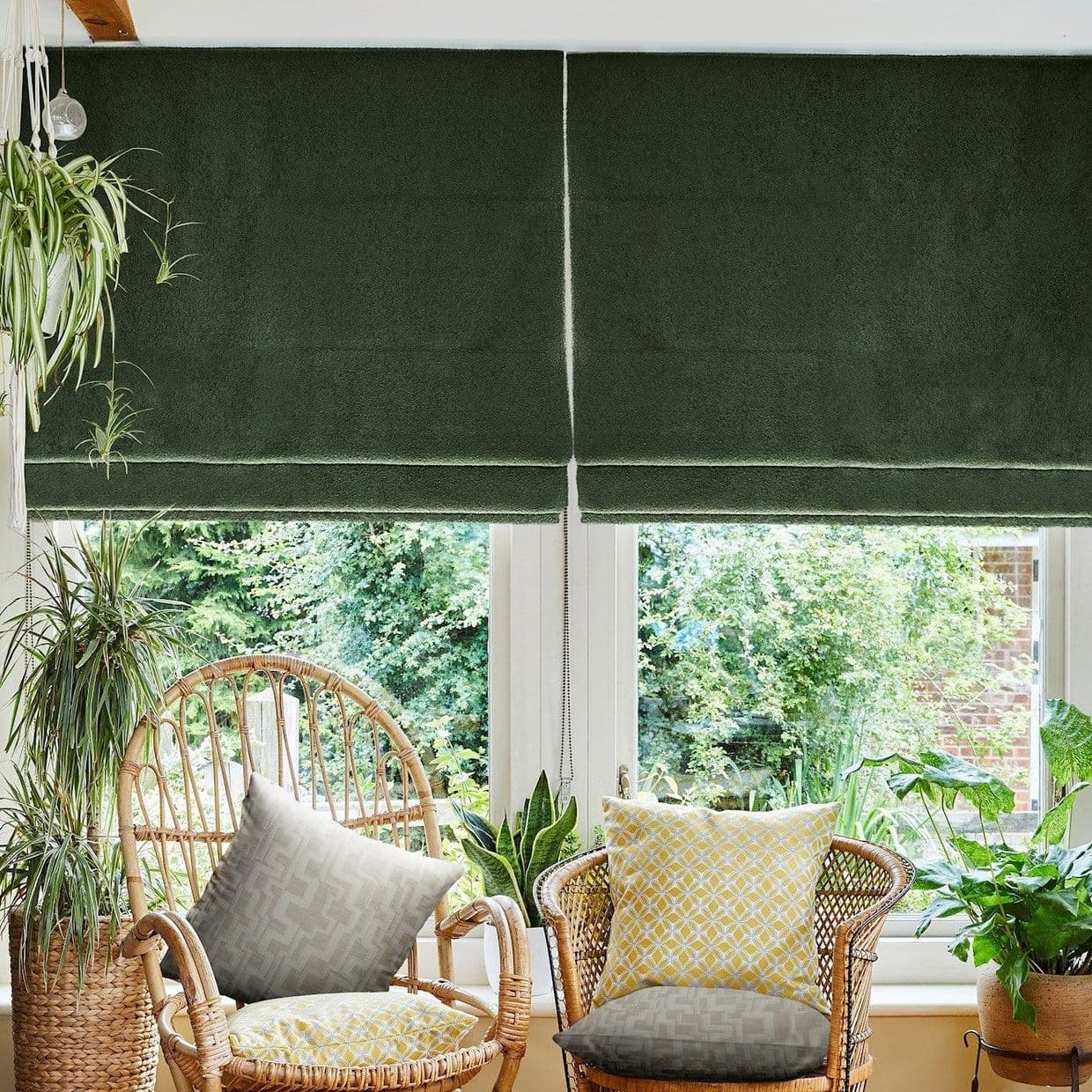 Deep green Roman blinds, lowered and split at the center with a visible pull chain, filtering light; in a sunlit seating nook with wicker chairs, patterned cushions, houseplants and garden view.