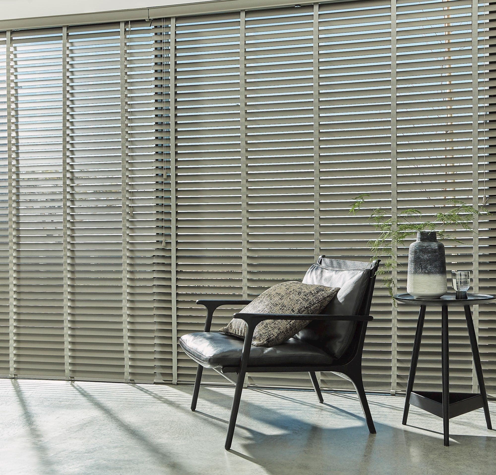 Horizontal Venetian blinds across a full-height window, slats tilted to filter light; casting striped shadows over a minimalist living corner with a leather armchair, side table and vase.