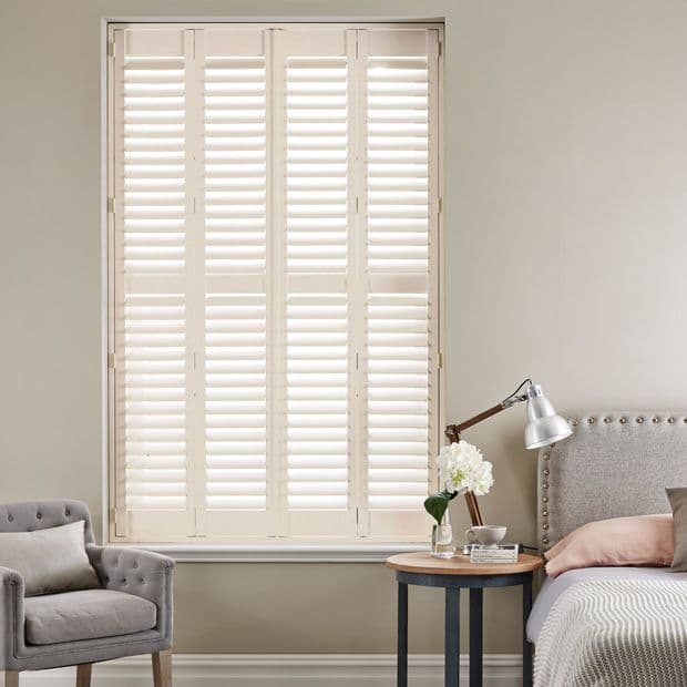 White plantation shutters covering a tall window, louvered panels closed and filtering soft daylight; set in a neutral-toned bedroom with upholstered bed, bedside table, lamp, and armchair.