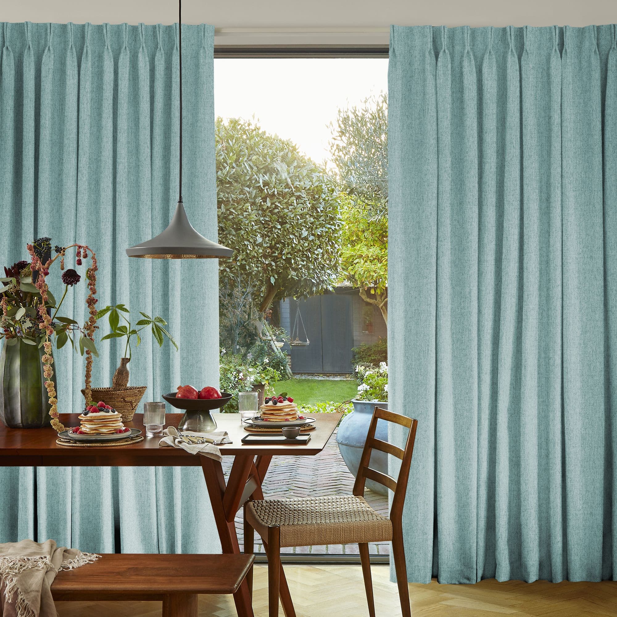 Pleated floor-to-ceiling turquoise curtains, parted at center in soft folds. They frame and filter sunlight. Dining table set for breakfast in bright dining room opening onto a green garden.