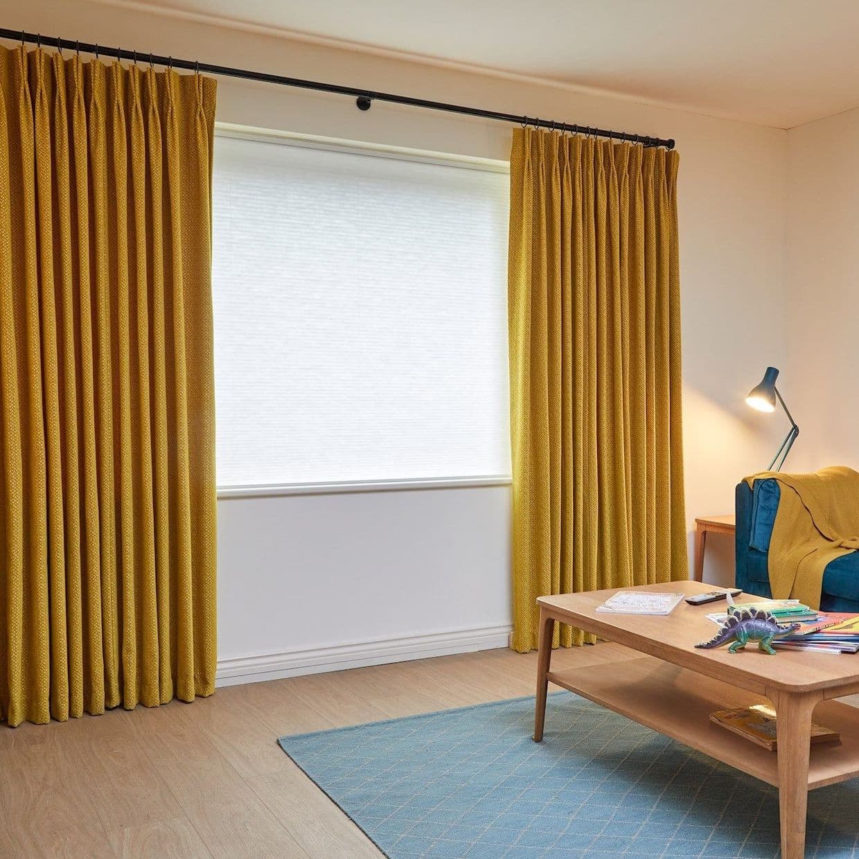 Mustard pleated curtains on a rod, parted to reveal closed white blind; curtains hang, filtering daylight in a living room with wooden floor, blue rug, coffee table and teal armchair.