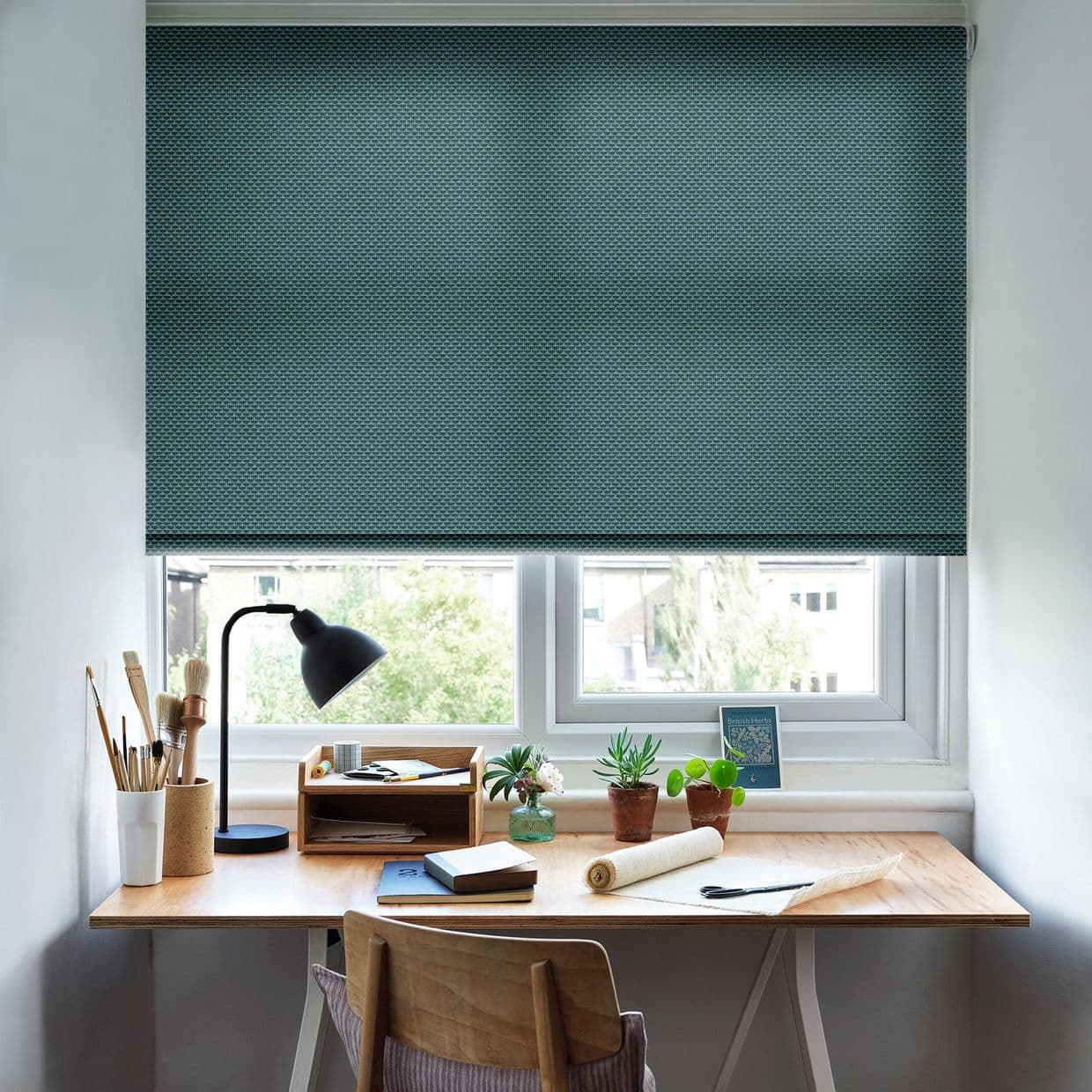 A teal, textured roller blind lowered almost fully across a double window, filtering light; shading a desk workspace with lamp, brushes, notebooks, small potted plants and a faint outdoor view.