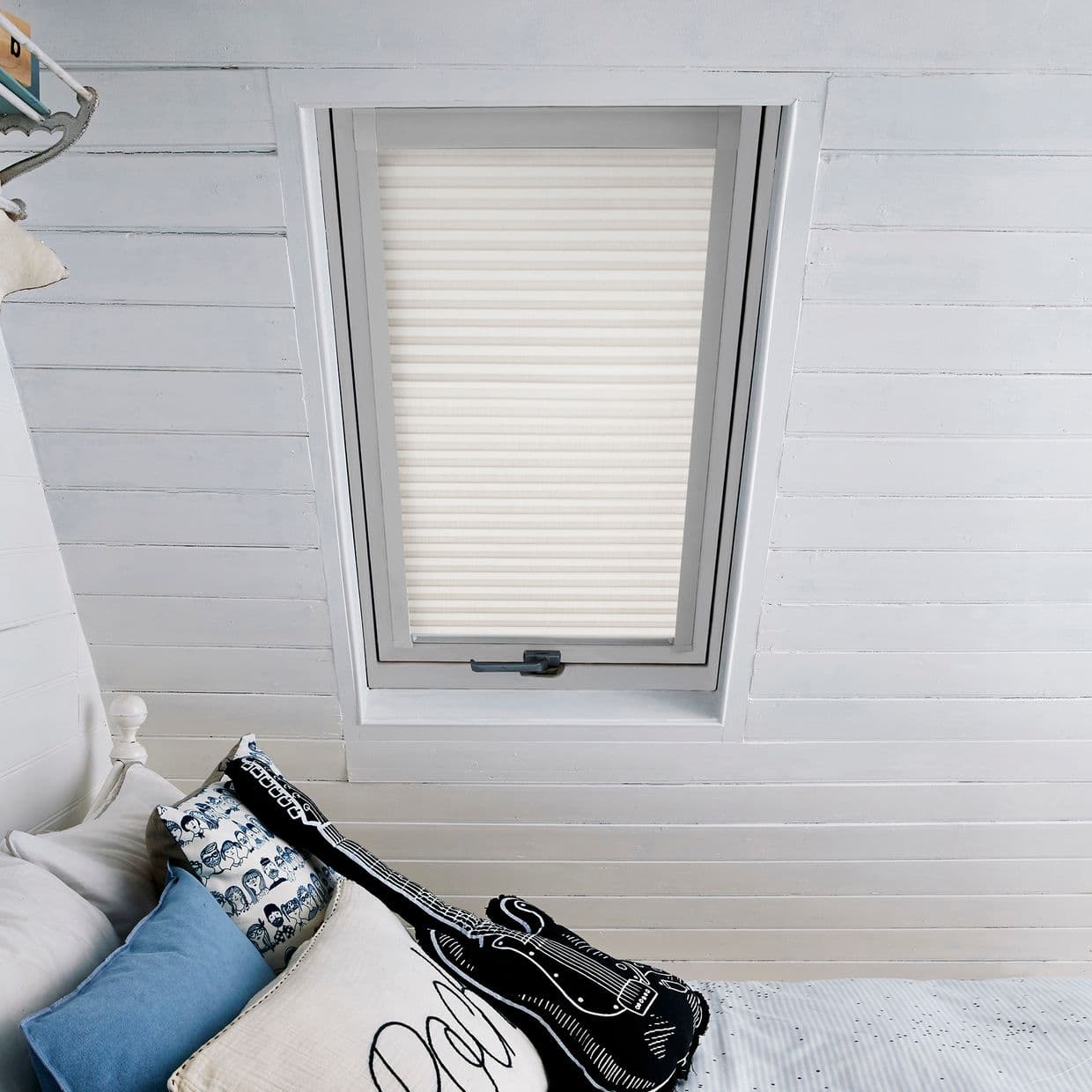 A cream-white pleated skylight blind, fully lowered within the window, diffuses soft daylight; in a sloped white wooden-ceiling attic bedroom above a bed with blue, patterned and guitar-shaped cushions.
