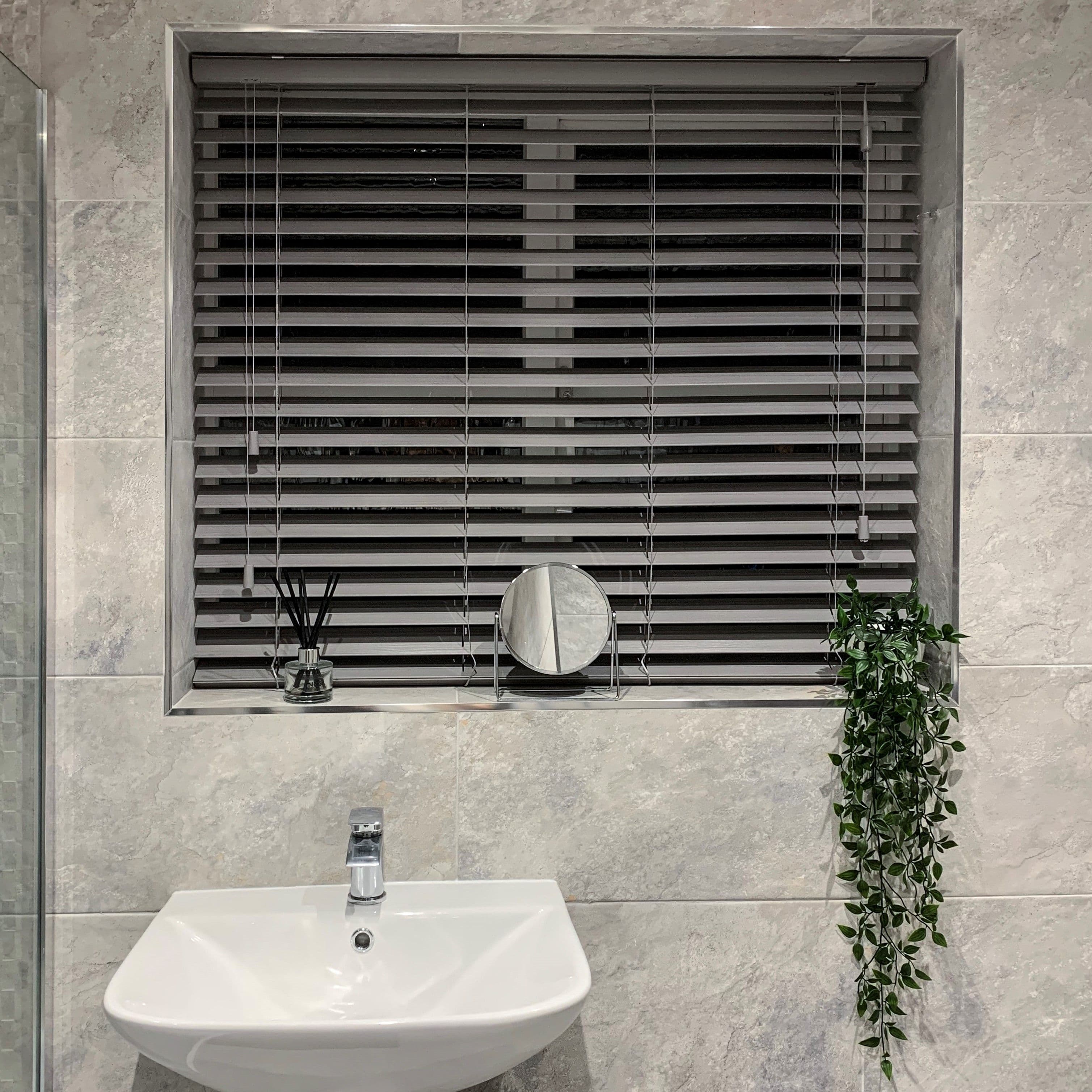 Grey horizontal venetian blinds, fully closed with visible cords and slats, filtering dim light through a recessed tiled bathroom window; white sink, small round mirror, diffuser, trailing plant on sill.