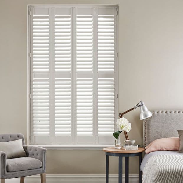 White plantation shutters covering a tall windowâ€”four framed panels with horizontal slats, closed and filtering soft daylightâ€”set in a neutral bedroom corner with upholstered headboard, bedside lamp, vase, and armchair.