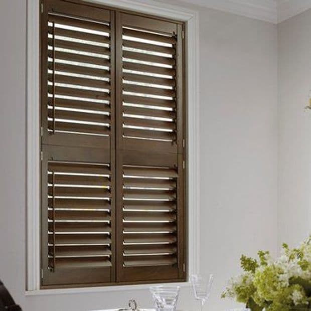 Brown wooden plantation shutters cover a tall window in two panels with slats; they are closed, angled to filter light, in a dining area with a vase of flowers.