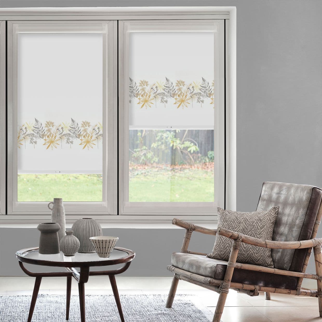 White roller blinds, lowered to mid-height across two windows and decorated with a gray-and-gold floral band, filter daylight into a neutral living room with a rattan chair and ceramics.