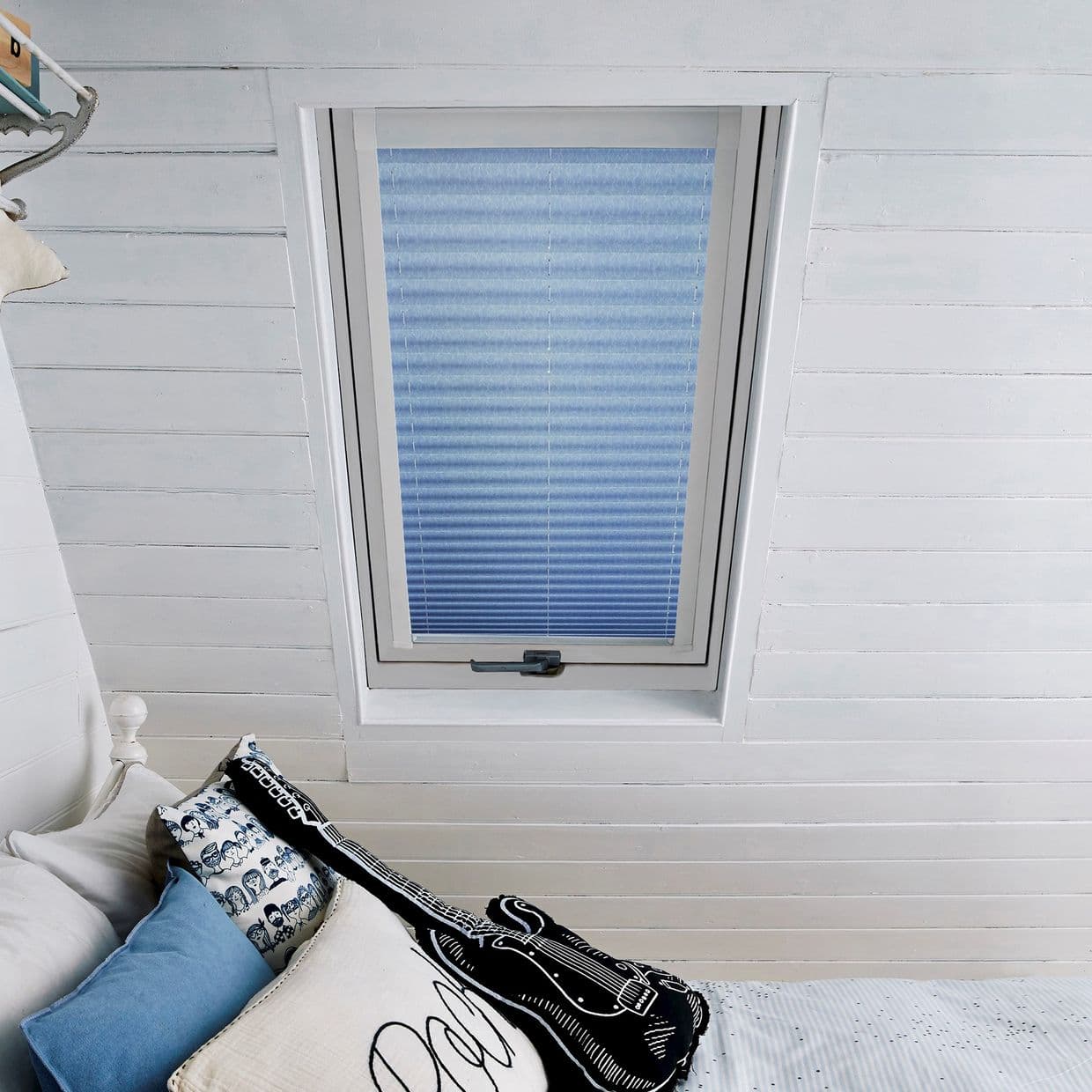 Blue pleated skylight blind centered in a white-framed roof window, fully lowered and filtering daylight; attic bedroom setting with white-painted tongue-and-groove walls and pillows, decorative guitar cushion.