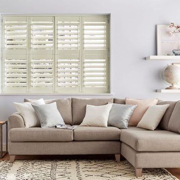 White plantation shutters covering a wide window, louvered slats tilted to filter soft daylight; set above a beige L-shaped sectional with pastel cushions in a bright, neutral living room.