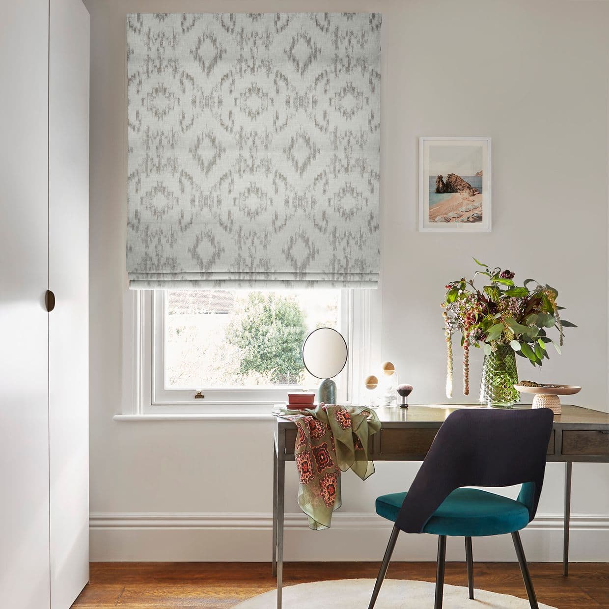 Patterned Roman blind (pale grey ikat) fully lowered over window, filtering soft daylight; small home office with wooden desk, mirror and vase of flowers, teal chair, neutral walls.