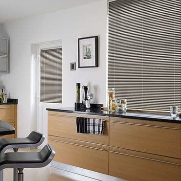 Horizontal beige-and-white Venetian blinds covering a large window and a glass door, closed and filtering daylight; modern kitchen with light wood cabinets, black countertop, bar stools, and minimalist decor.