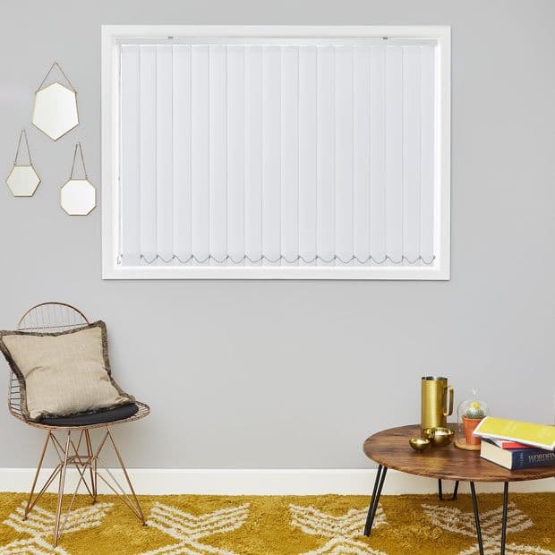 Vertical white blinds covering a window, closed and filtering daylight; mounted on a gray living-room wall above a mustard patterned rug, wire chair with cushion and wooden coffee table.