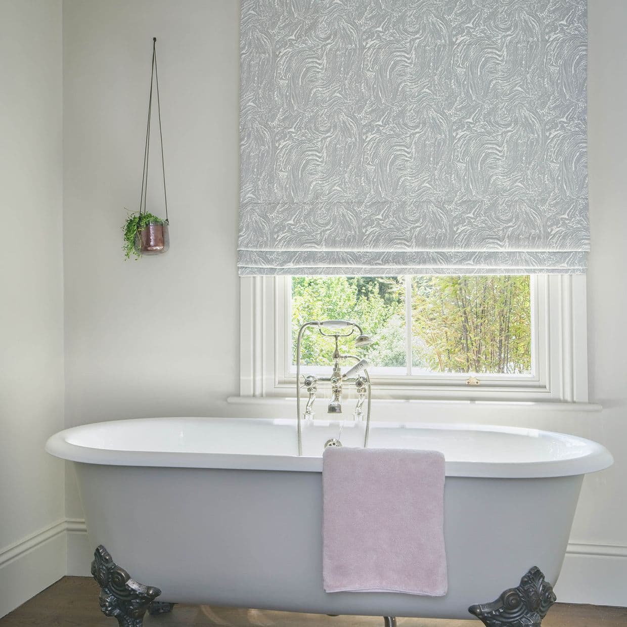 Patterned grey Roman blind, partially lowered with swirling texture, filtering daylight over a window above a freestanding claw-foot bathtub with chrome faucet, pink towel and hanging plant.