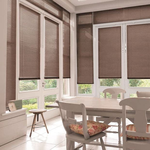 Brown horizontal slatted blinds covering tall white-framed bay windows, partially lowered, filtering soft daylight; dining table and chairs in a bright breakfast nook overlooking a green garden.