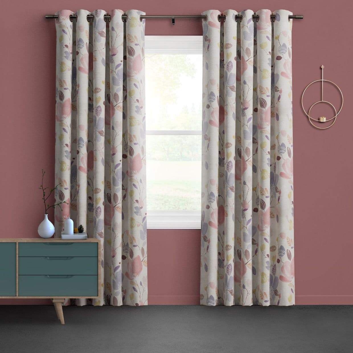 Grommet-top, full-length floral-leaf curtains, parted at center to diffuse bright daylight through a white-framed window, against a mauve wall with a teal sideboard and circular wall candle holder.