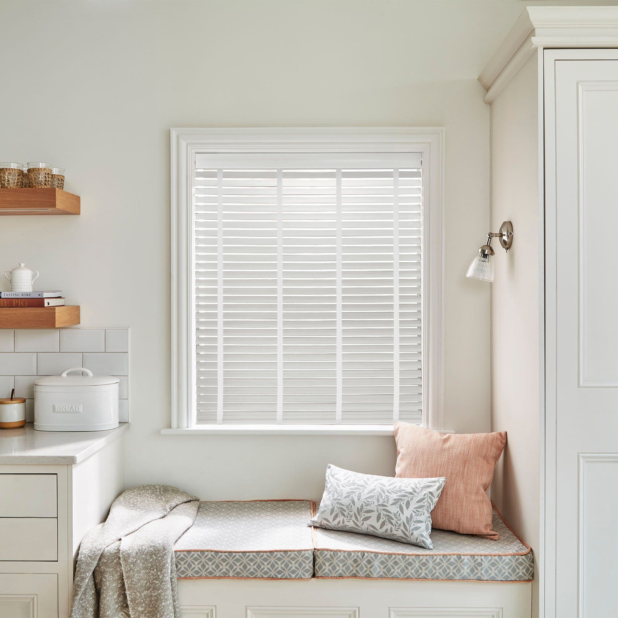 White horizontal slatted blinds fitted inside the window frame, fully closed and diffusing soft daylight; below, a cushioned bench nook with patterned cushions, wall sconce, and open shelves. BREAD