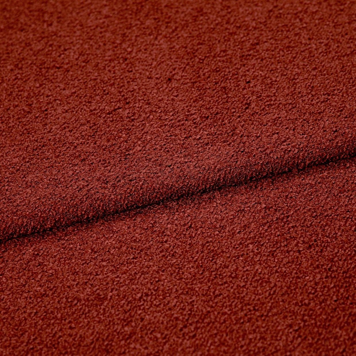 Rust-red textured boucle fabric folded across the frame, creating a dark seam; close-up studio view highlighting coarse looped surface and even, warm lighting.