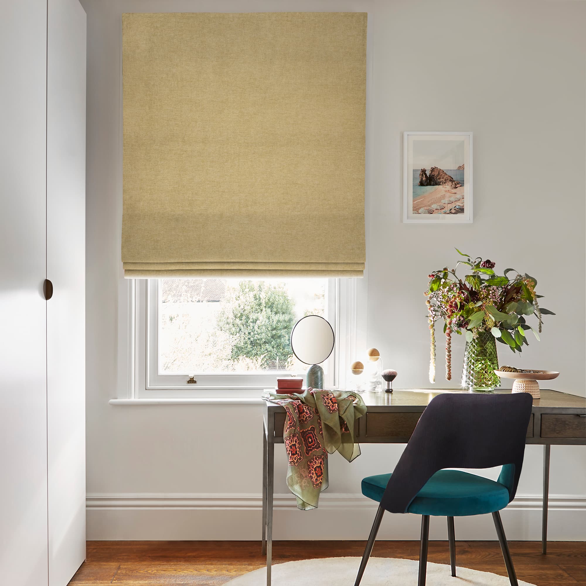 Beige Roman blind, lowered with soft folds and textured fabric, filtering daylight through the window; home workspace beneath with a desk, teal chair, vase of flowers and framed seaside print.