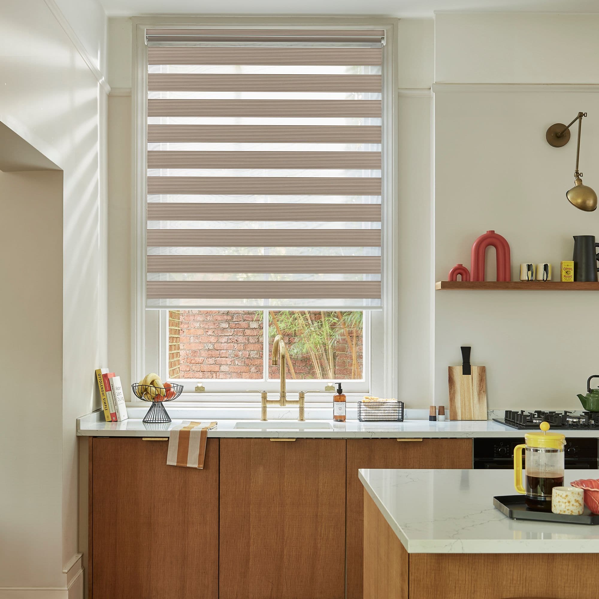 Layered zebra roller blind, lowered with alternating opaque and sheer horizontal bands, filtering soft daylight; modern kitchen setting with white walls, wood cabinets, brass faucet and countertop island.
