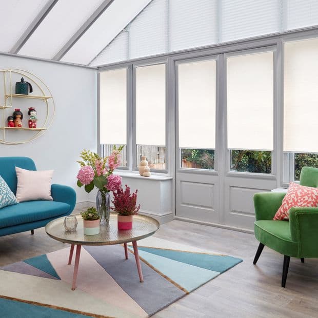 Cream pleated cellular blinds cover tall conservatory windows and doors, mostly lowered and diffusing bright daylight; a coffee table, turquoise sofa, green armchair and flowers on a pastel geometric rug.