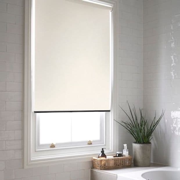 Beige roller blind with thin black hem fully lowered over a window, blocking light and revealing lower panes; a bright tiled bathroom, sill holds a potted plant and wicker tray.