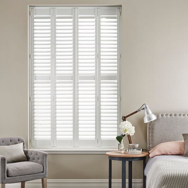 White plantation shutters covering a floor-to-ceiling window; slatted panels closed, filtering soft daylight. Context: neutral-toned bedroom nook with upholstered armchair, bedside table, lamp and floral vase.