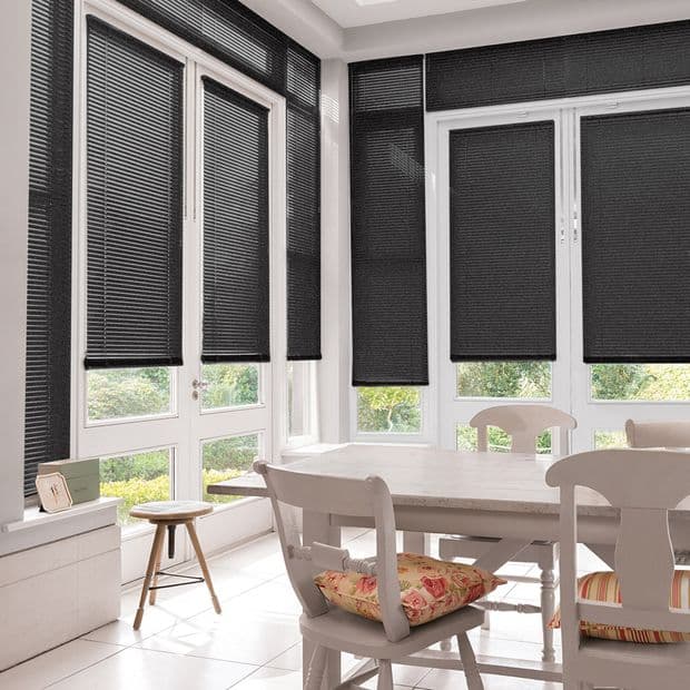 Black pleated cellular blinds, lowered mostly to mid-height with narrow raised gaps, filtering daylight and blocking view; dining room with white table and chairs, tiled floor and garden beyond.
