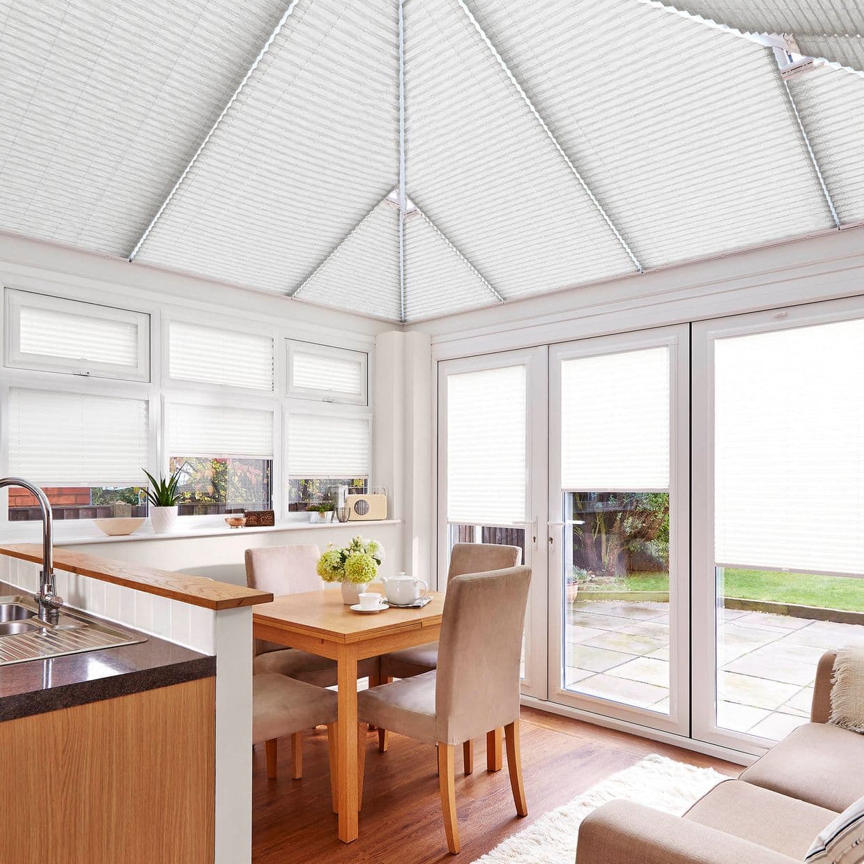 White pleated cellular blinds cover conservatory roof panels, upper transom windows, and glass patio doors, filtering bright daylight into a small kitchen-dining area with wooden table, chairs, and garden view.