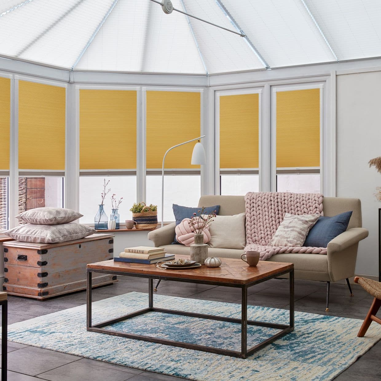 Honeycomb pleated blinds lowered to mid-height across conservatory windows, filtering warm light; beige sofa with chunky knitted throw and pillows, wooden coffee table on a blue rug in a bright living room.