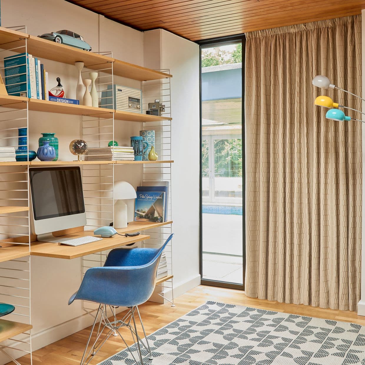 Beige pleated floor-to-ceiling curtains drawn closed, softly filtering daylight; they cover a tall glass door beside a wooden-shelved home office with a desk, blue chair, computer, patterned rug.

DESIGN
Palm Springs Weekend