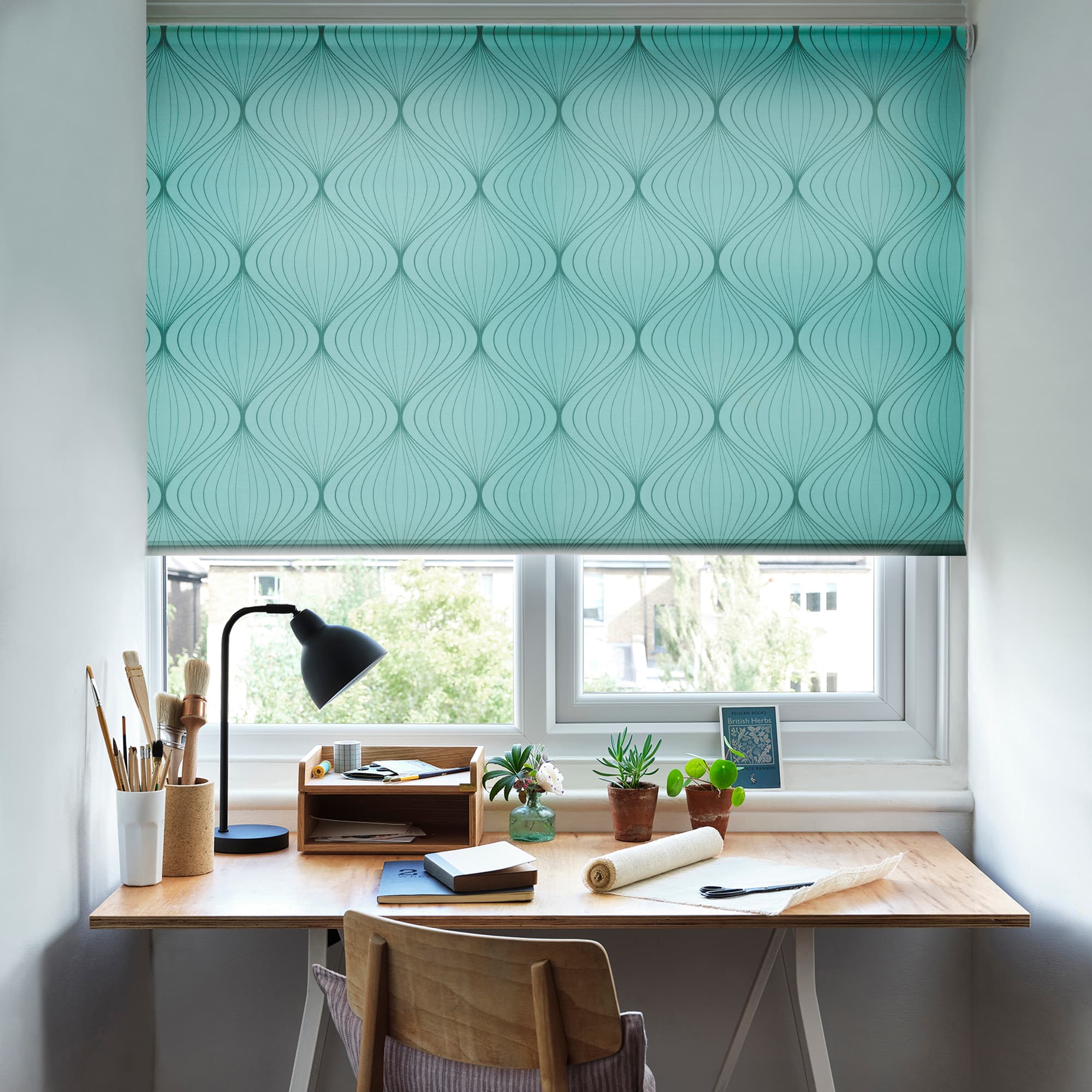 Teal patterned roller blind (repeating curved teardrop motif) lowered over the window, filtering daylight; home desk workspace beneath with lamp, art supplies, small potted plants on the windowsill and trees outside.