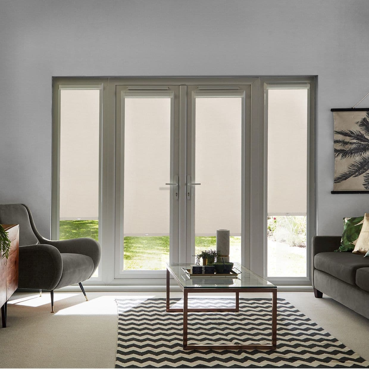 Beige roller blinds covering a set of central French doors and sidelights, fully lowered with short lower gaps; filtering daylight into a living room with grey seating and garden view.