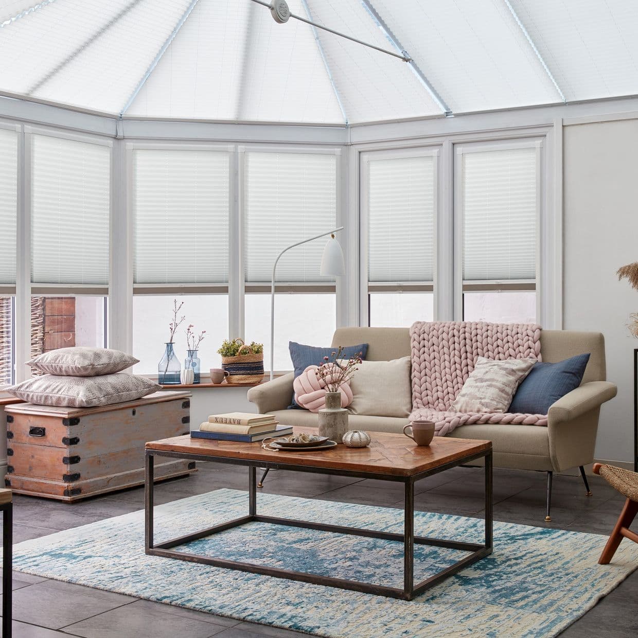 White pleated cellular blinds cover bay windows and the conservatory roof, drawn to filter soft daylight; cozy living room with beige sofa, wooden coffee table, pastel throw and decorative vases.