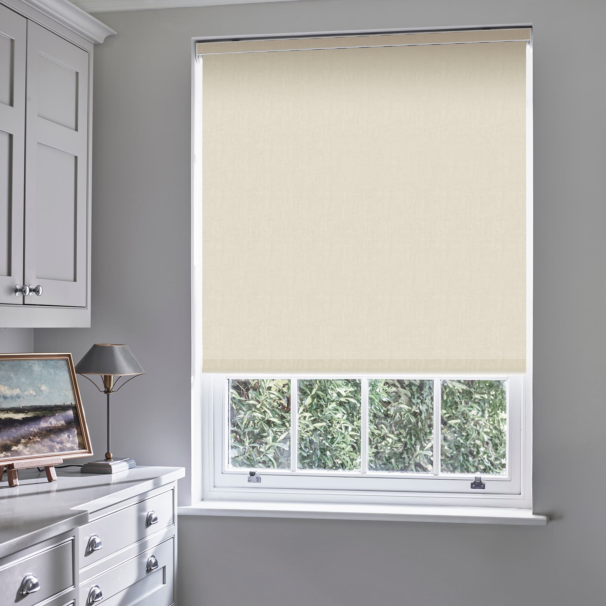 Beige fabric roller blind, fully lowered, subtly textured and light-filtering; covering a white sash window in a softly lit grey room with white cabinetry, dresser, lamp and leafy garden view.