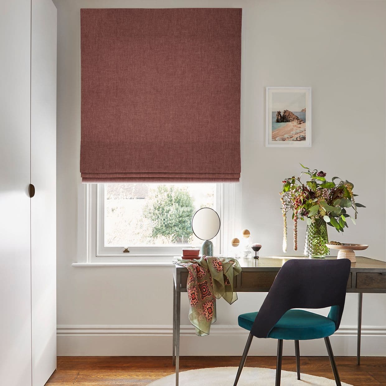 Roman fabric blind, dusty-rose textured with soft folds, lowered nearly fully over the window. Filtering daylight through a narrow bottom gap, above a desk with mirror, chair and vase.
