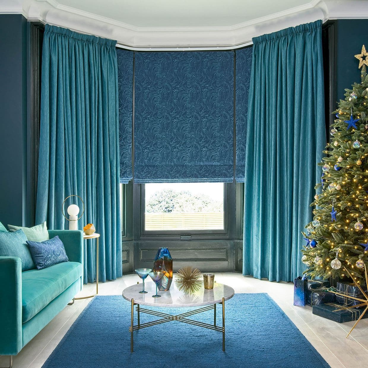 Teal velvet full-length curtains frame a bay window with a patterned roman blind partially lowered, filtering daylight; contemporary living room with teal sofa, marble coffee table and decorated Christmas tree.