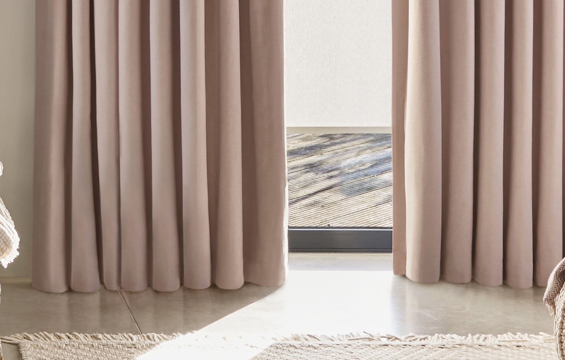 Floor-length beige pleated curtains, hanging mostly closed with a narrow central gap, filtering daylight; a white roller blind and outside wooden deck visible beyond; neutral living-room concrete floor and rug.