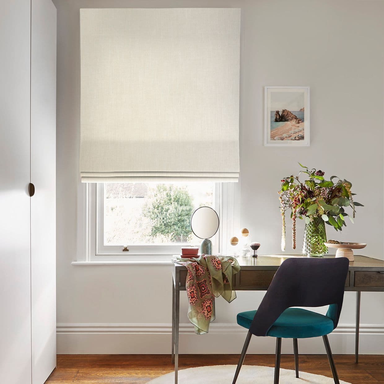 A beige fabric Roman blind, lowered with neat folds, filtering soft daylight through the window above a desk in a neutral home office with a teal chair and vase.